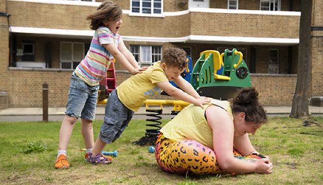 A woman sits on the floor, stretching, while her two children help her by pushing on her back.
