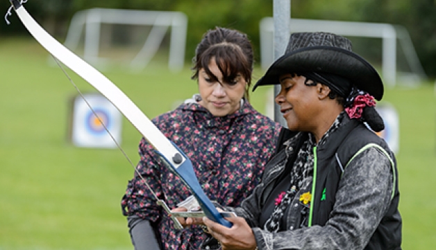 A female archery coach instructs another woman at an archery lesson outdoors.