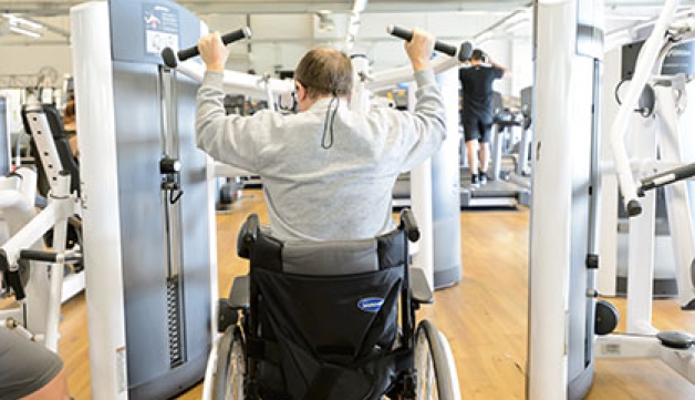 A man in a wheelchair uses a lat pull down machine in a gym.