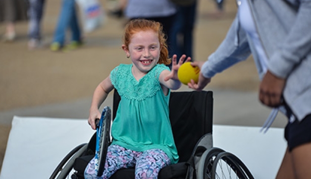 A girl in a wheelchair takes a tennis ball from a coach 