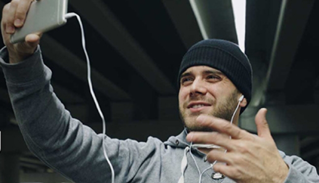 A man in a tracksuit top talks to his phone, while wearing headphones, delivering a sports session online.