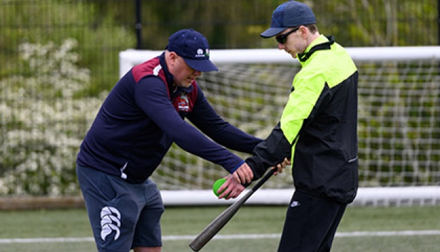 An instructor assists an visually impaired man to hit a tennis ball with a softball bat.