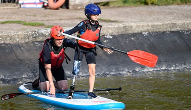 A boy with a prosthetic leg paddles a paddle board, with an adult assistant on the back of it.