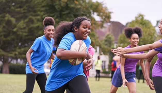 A girl runs with a rugby ball as a group of girls play rugby in a park.
