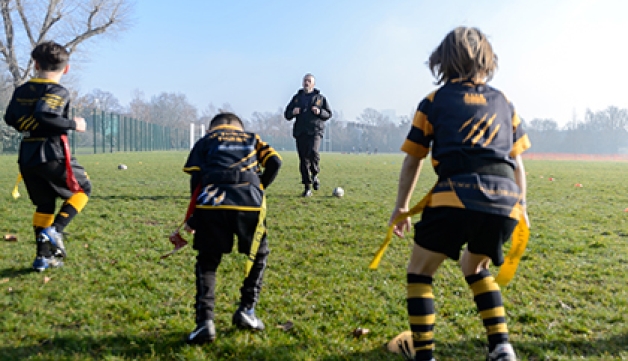 A coach instructs three young boys how to play tag rugby, on an open playing field