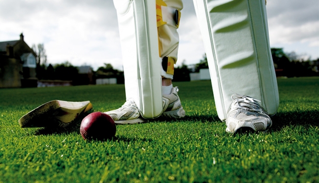 A cricket bat and ball on the grass with a cricketer's legs in the background