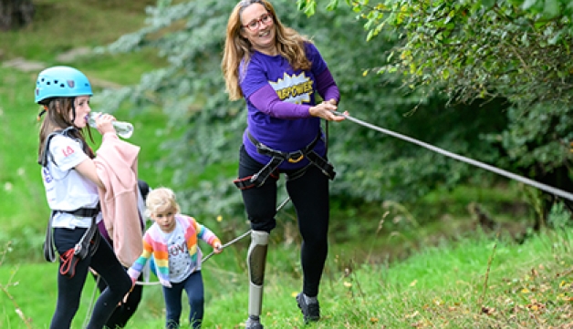 A woman with a prosthetic leg uses a rope to climb up a slope and two small girls watch on.