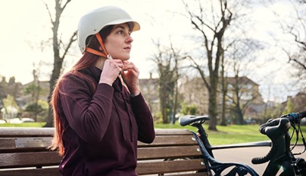 A woman, sitting on a bench in a park, fastens her cycling helmet as her bike leans up against the end of the bench.
