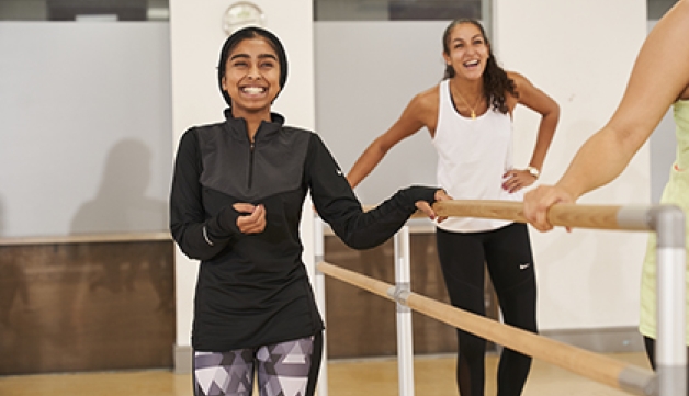 A smiling woman stands with one hand on a ballet barre, while another in the background also smiles and laughs