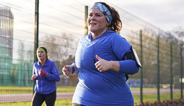 A woman, wearing headphones, runs in a park, with another runner in the background