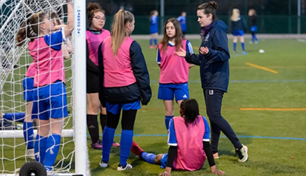 A female coach speaks to a teenage girls' football team at the side of an artificial football pitch