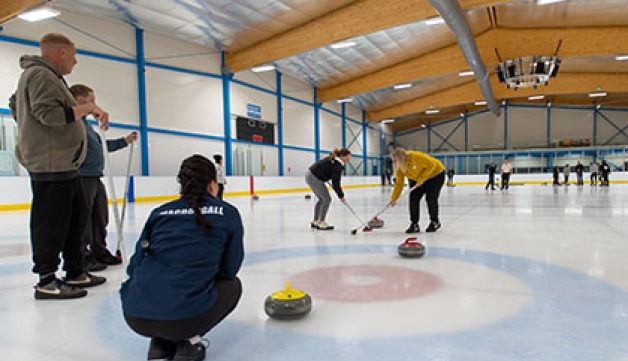 Two women sweep a curling stone as another crouches in the 'house' and two men watch on, also standing on the ice