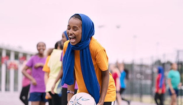 A girl wearing a head scarf laughs and smiles while playing netball in a school playground