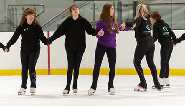 Some young girls and boy skate holding each other arms and making a big circle on an indoors ice rink.