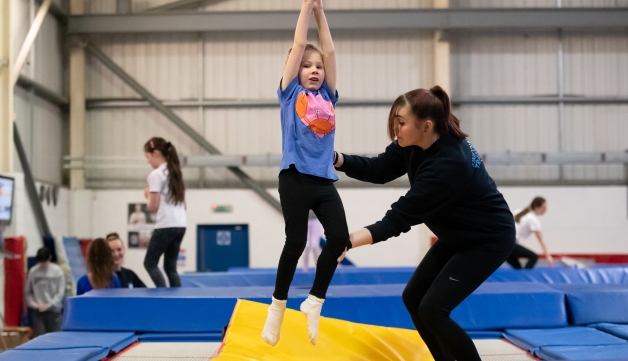 A coach helps a little girl keep straight and she jumps on a trampoline on an indoors club.