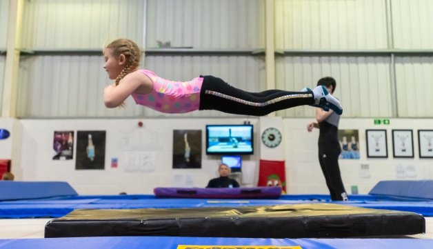 A girl extends her body in the air as she jumps on a trampoline and next to her an older girl is about to jump on another trampoline while a coach observes from the floor of an indoors club.