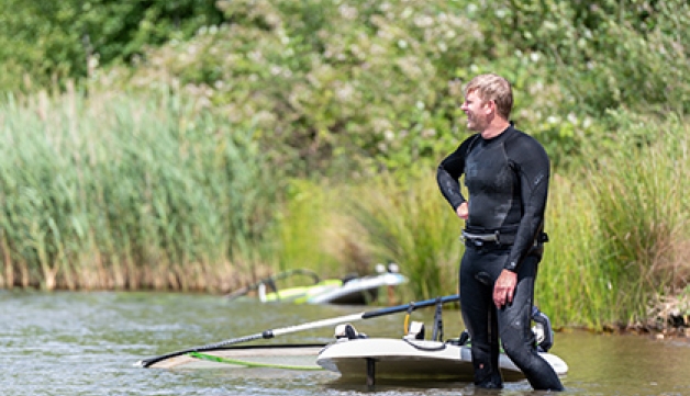 A man in a wetsuit stands in shin-deep water in a lake, next to a windsurfing board and sail
