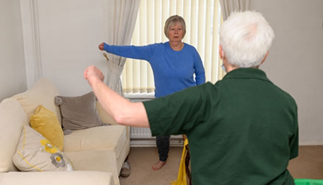 An elderly woman follows directions from a healthcare professional to stretch her right arm on her living room. 