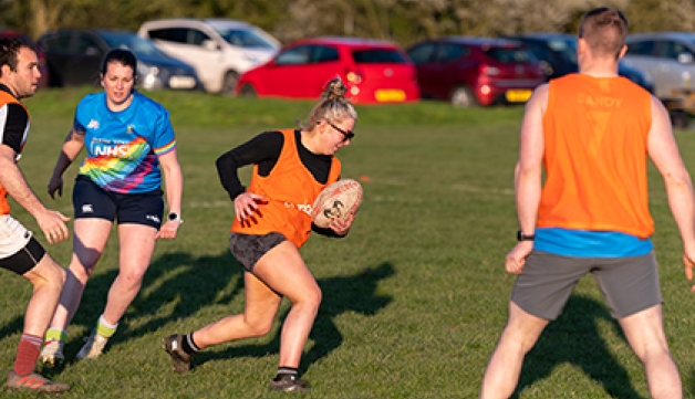 A woman runs as players from both teams look at her during an outdoors rugby game.