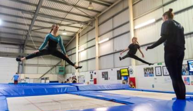 A volunteer gives instructions to two young girls who are bouncing on trampolines, their legs are stretched out wide