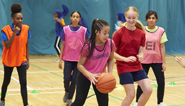 A young person holding a basketball on a court is surrounded by four other players