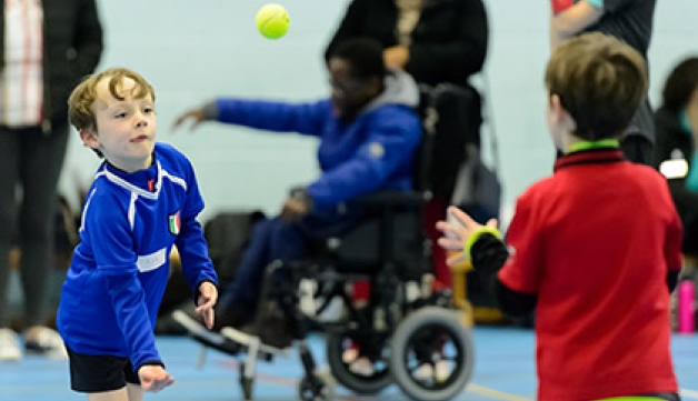 A child throws a tennis ball to another child. There is a wheelchair user in the background.