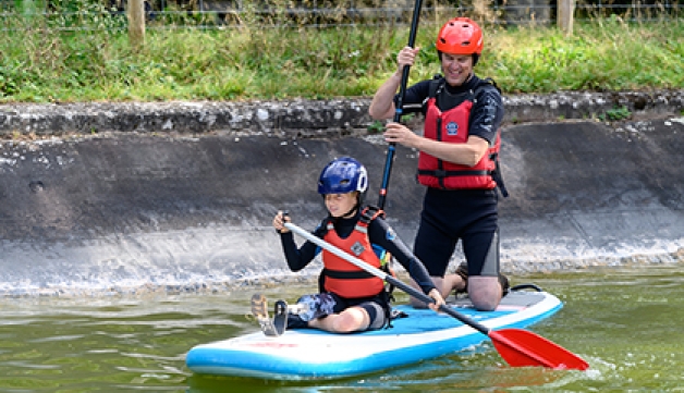 A boy with a prosthetic leg stands on a paddle board, on water. while an instructor kneels behind him