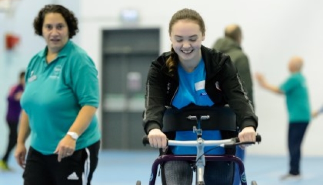 A young person smiles while walking with a wheeled aid in a sports hall, while a coach watches on