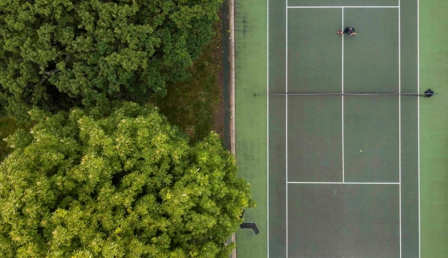 An overhead shot of a green tennis court in a park, next to green trees.