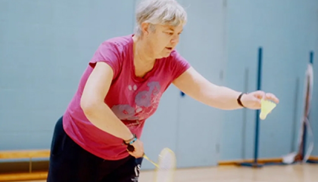 An older woman prepares to serve while playing badminton in a sports hall