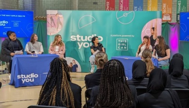 A group of secondary school students sat on chairs in a sports hall watch a panel discussion between a host and five women.