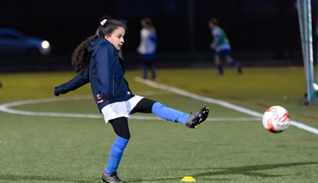 A girl kicks a football on an outdoors pitch.