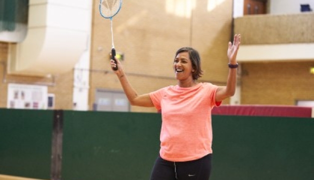 A smiling woman raises both arms, one of them holding a badminton racquet, on a court in a sports hall.