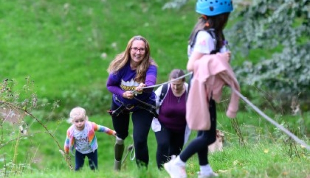 A woman smiles while pulling a rope to climb a hill. A girl in a helmet is at the top of the hill and there are other climbers below.