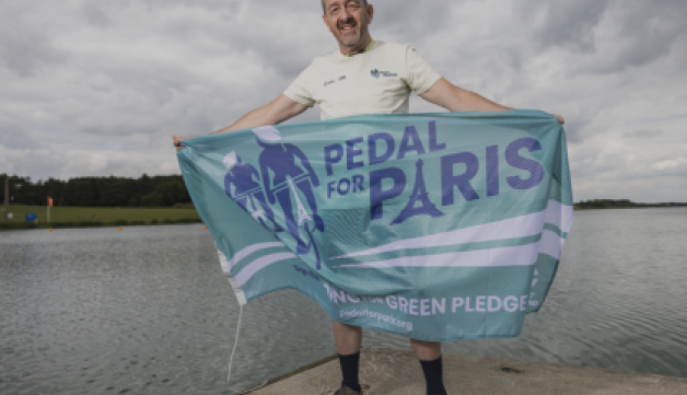 Chris Boardman holds a Pedal for Paris flag in front of a lake