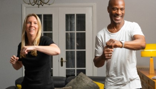 A smiling man and a woman stood side by side in a living room, taking part in a workout.