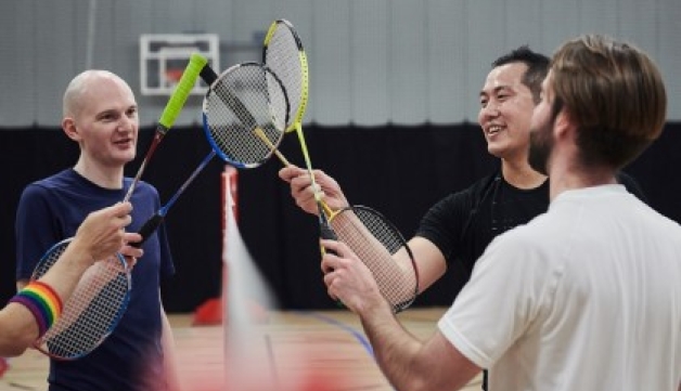 Four people touch their badminton racquets together over the net after a game on an indoor court
