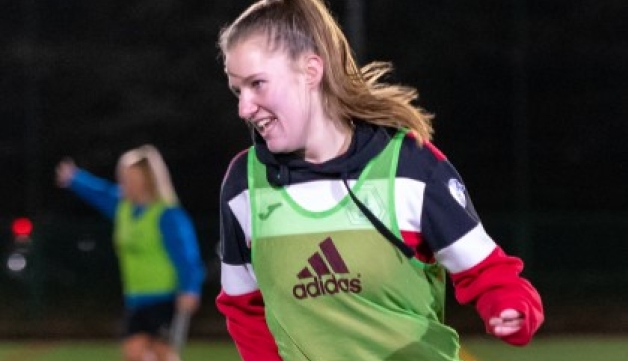 A young woman in a football bib smiles as she moves on an outdoor artificial pitch at night.