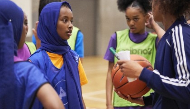 A female coach holding a basketball, pen and paper talks to a group of girls on a court.