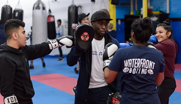 Three young people in a boxing gym gather round their coach as he demonstrates using boxing pads