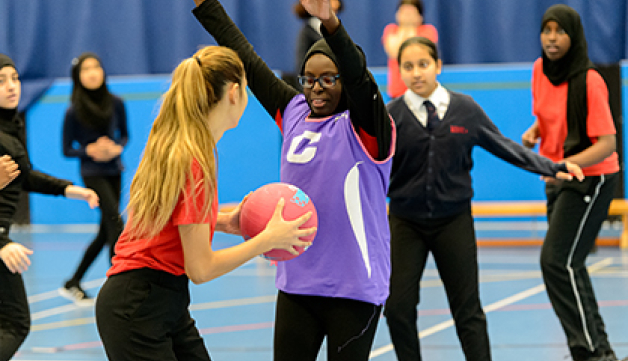 Girls play netball in a PE lesson. One player has the ball and another girl is guarding her, with both arms in the air.