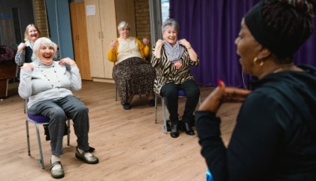 Four older women touch their shoulders while taking part in a seated exercise session with a trainer in a hall.