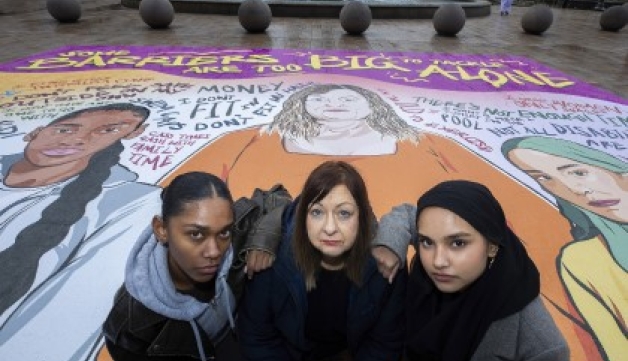 Three women stand on a giant mural featuring illustrations of women and the text 'Some barriers are too big to tackle alone'.