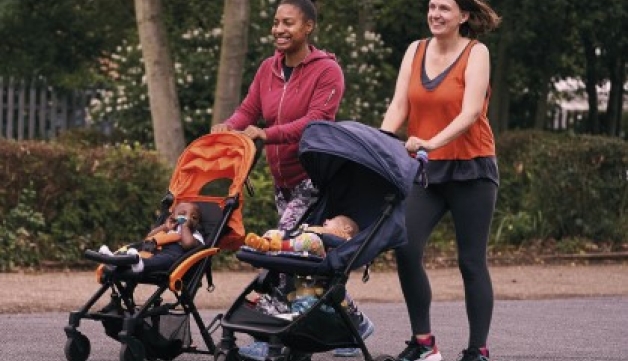 Two women in athleisure clothing push their children in prams on a path.