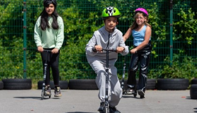 Three children wearing helmets ride on scooters.