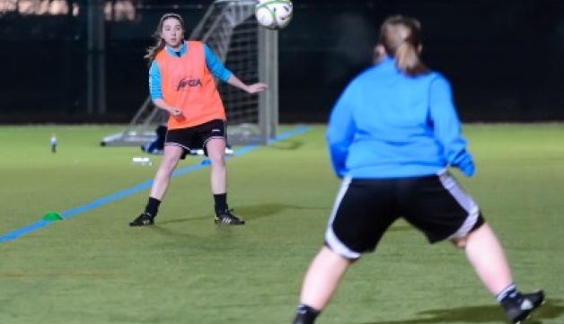 Two young female footballers with the ball in the air on an outdoor artificial grass pitch at night.
