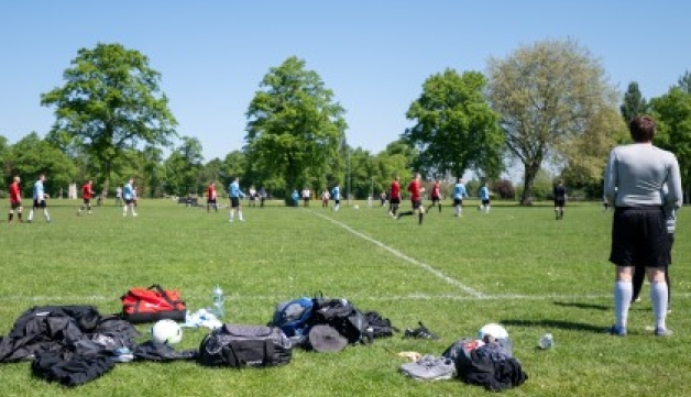 A football coach stands beside equipment bags on the sideline of a football match on a playing field on a summer's day, with mature trees in the background.