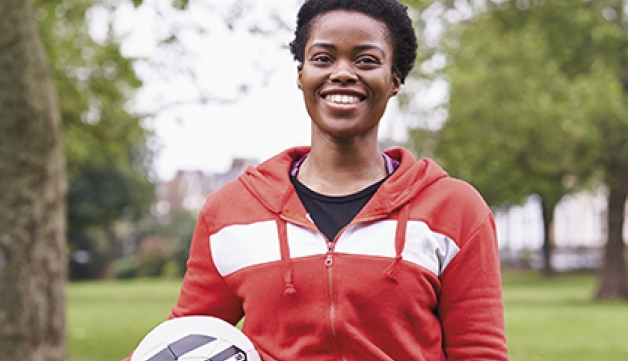A black young woman holds a football while standing in a park or a cloudy day.