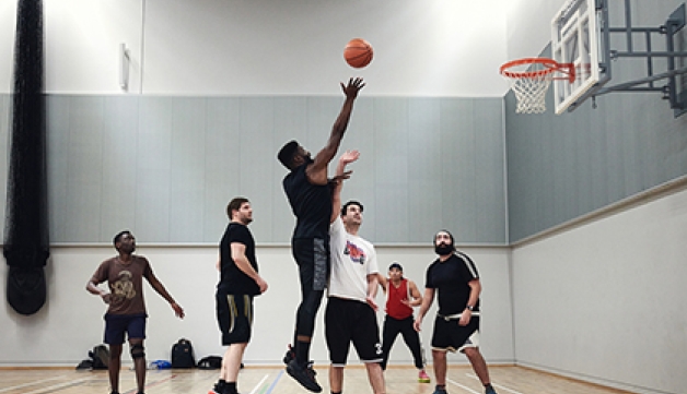 A man shoots a basketball over another player who tries to block it, while other players watch on
