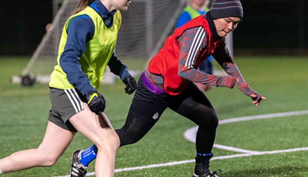 Women wearing training bibs of different colours play football during an evening training session outdoors.